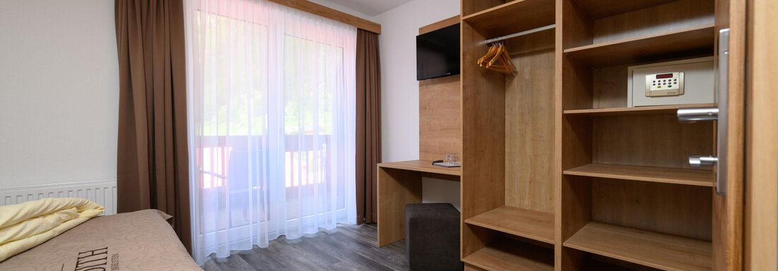A bright room with a large window and a desk. There is a wardrobe and a cozy corner with a bed. - Gästehaus Judith - Zwieselstein