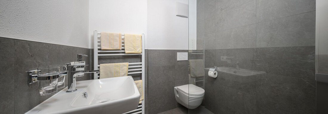 A modern bathroom with gray tiles and a sink. There is a toilet and a towel rack with fresh towels. - Gästehaus Judith - Zwieselstein