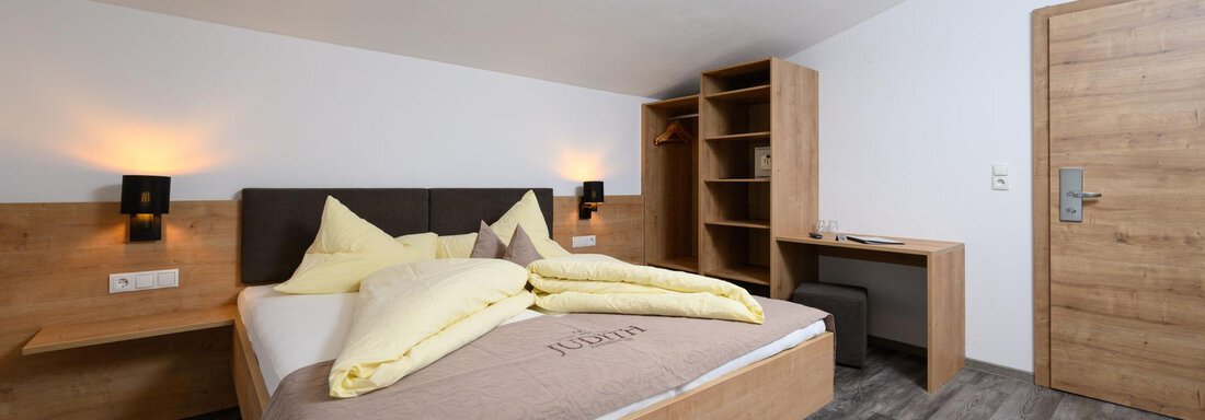 A modern bedroom with a double bed, wooden furniture, and a table. The walls are bright, and there is pleasant lighting. - Gästehaus Judith - Zwieselstein