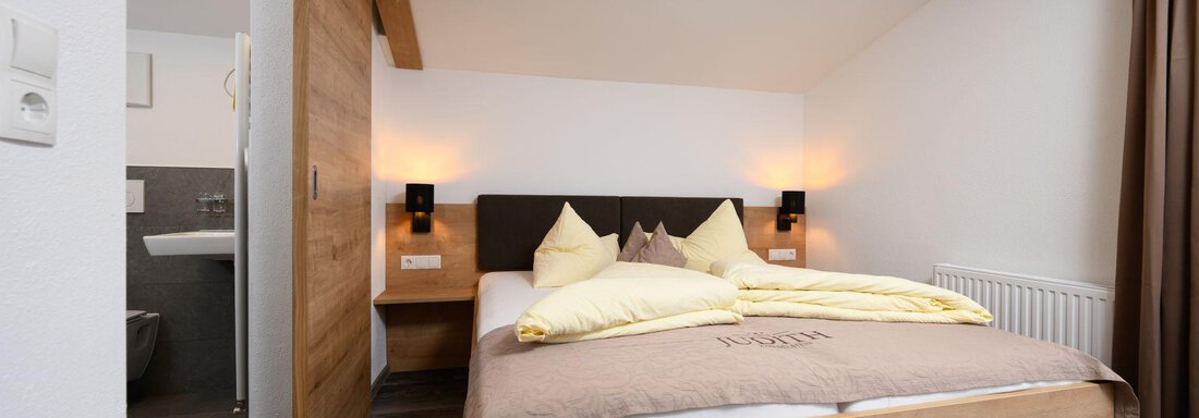 A cozy bedroom with a large bed and wooden details. There is soft lighting and access to the bathroom. - Gästehaus Judith - Zwieselstein