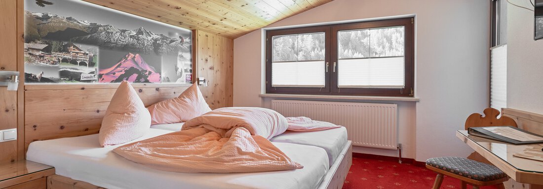 Doppelzimmer A cozy bedroom with a double bed and wooden paneling. The room has large windows and a red carpet. - Gästehaus Judith - Zwieselstein