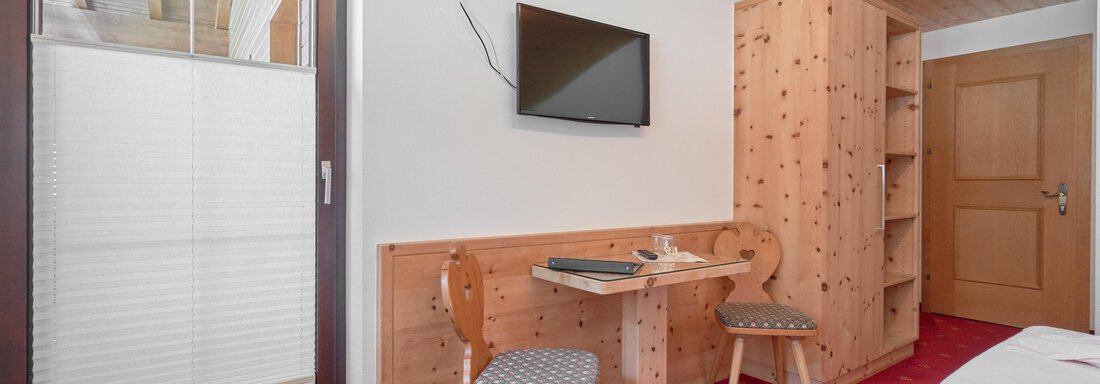 A cozy room with wooden furniture and a television on the wall. The room is simply furnished, with a table and two chairs. - Gästehaus Judith - Zwieselstein