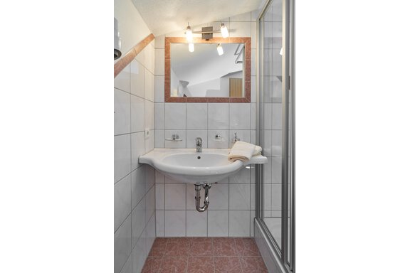 A modern bathroom with a white tiled wall and a sink. In the room, a mirror and a shower cabin are visible. - Gästehaus Judith - Zwieselstein