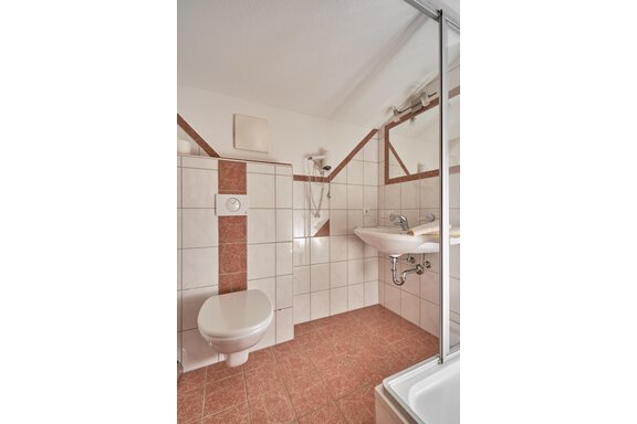 A modern bathroom with white tiles and brown accents. There is a shower, a sink, and a toilet. - Gästehaus Judith - Zwieselstein
