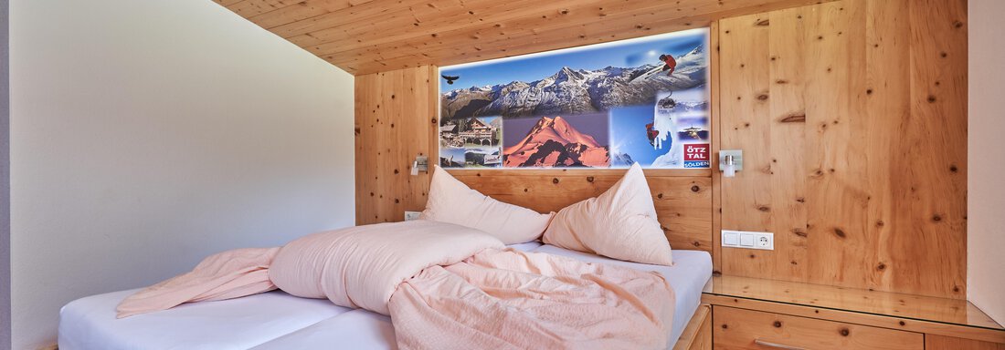 A cozy bedroom with a wooden frame bed and two pillows. On the wall hang pictures of mountains and activities in nature. - Gästehaus Judith - Zwieselstein