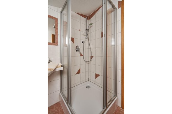 A modern shower cabin with glass walls and a handheld shower. The walls are tiled, and there is a towel on a shelf. - Gästehaus Judith - Zwieselstein