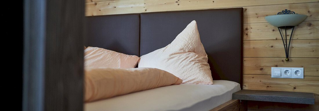 Schlafzimmer Appartement Nr. 1 A cozy bedroom with a wooden bed and large pillows. The walls are made of wood, and a wall lamp provides soft light. - Gästehaus Judith - Zwieselstein