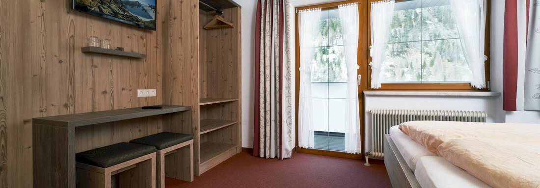 A cozy room with wooden furniture and a TV on the wall. The large windows provide a view outside and let in plenty of light. - Gästehaus Judith - Zwieselstein