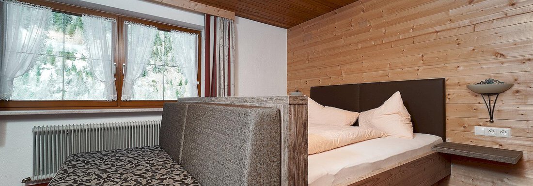 A cozy bedroom with wood paneling and large windows. There is a double bed and a sofa bed that look inviting. - Gästehaus Judith - Zwieselstein