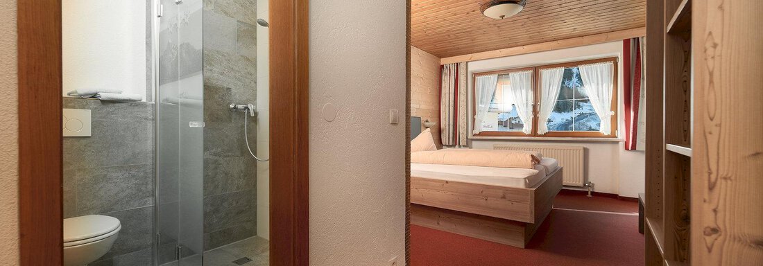 A cozy bedroom with a large bed and a bathroom with a shower. The room has wooden paneling and large windows for daylight. - Gästehaus Judith - Zwieselstein