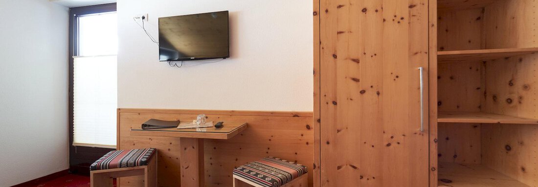 A simple room with wooden furniture and a TV on the wall. In the room, there is a table with two chairs and a wardrobe. - Gästehaus Judith - Zwieselstein