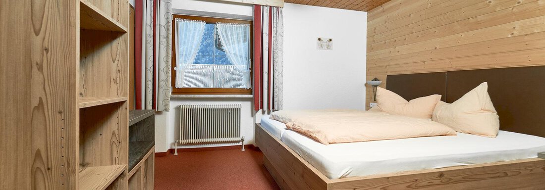 A cozy bedroom with wooden decor and a large window. The room includes a comfortable bed and an open wardrobe. - Gästehaus Judith - Zwieselstein
