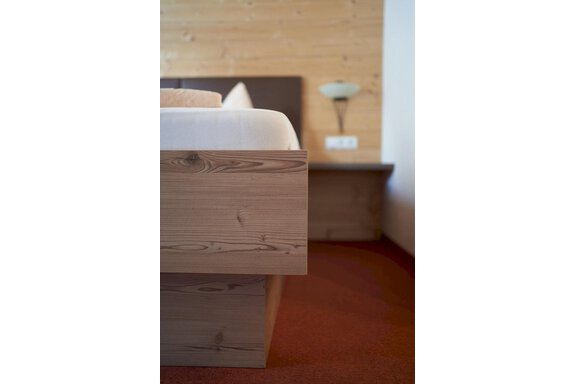 A cozy bedroom with a wooden bed and a warm, natural atmosphere. The wall is made of wood, and a nightstand with a lamp is visible in the background. - Gästehaus Judith - Zwieselstein