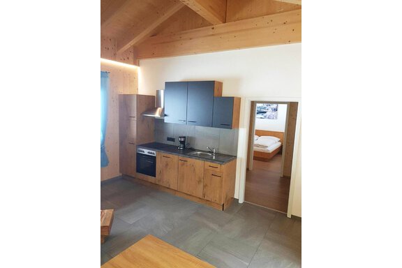 A modern kitchen unit made of wood with electrical appliances. In the background, a bedroom is visible. - Haus Fortuna - Zwieselstein
