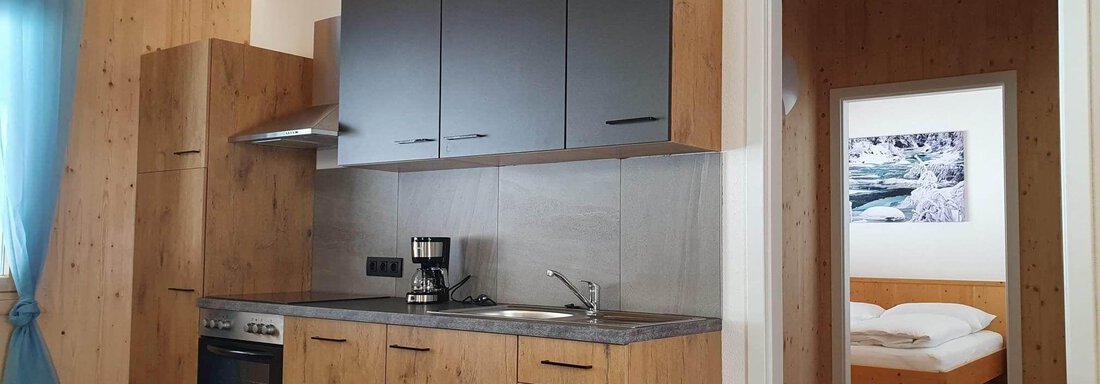 A modern kitchen with wooden cabinets and grey fronts. In the background, a sleeping area with a bed is visible. - Haus Fortuna - Zwieselstein