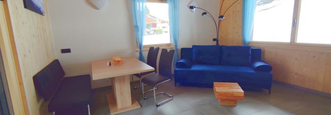 A modern living and dining room with a blue sofa, a wooden table, and elegant chairs. Large windows with light blue curtains let in a lot of light. - Haus Fortuna - Zwieselstein