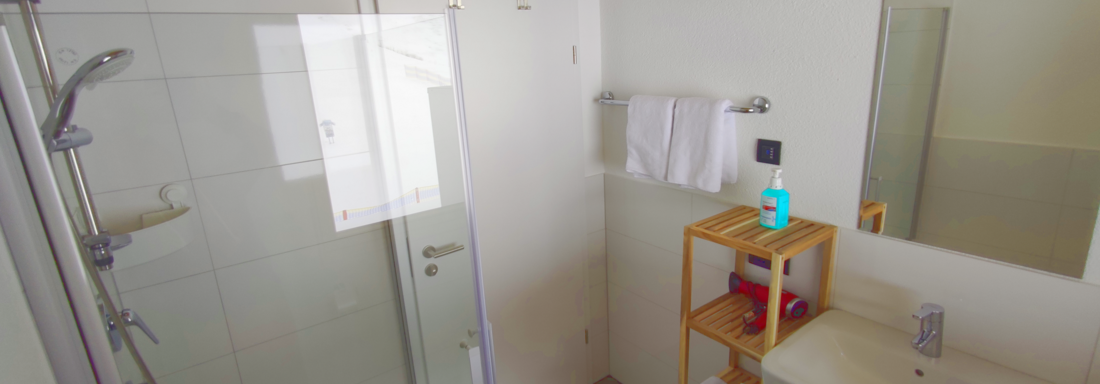 A modern bathroom with a shower and a sink. There are towels and toiletries on a wooden shelf. - Haus Fortuna - Zwieselstein