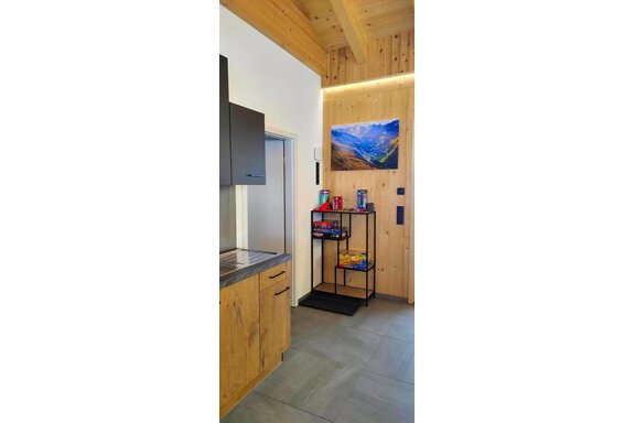 A modern kitchen with wooden cabinets and a view of a mountain. In the background, there is a shelf with snacks and drinks. - Haus Fortuna - Zwieselstein