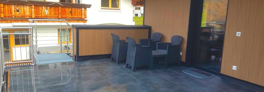 A modern terrace with black tiles and a plastic dining table. In the background, wooden balconies and a window can be seen. - Haus Fortuna - Zwieselstein