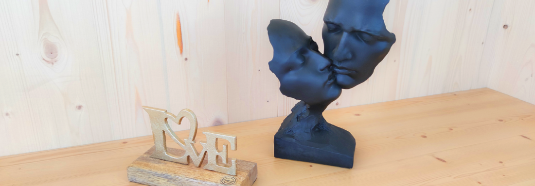 A modern sculpture of two faces kissing stands next to a wooden base with the inscription "LOVE". The background consists of light wood, creating a cozy atmosphere. - Haus Fortuna - Zwieselstein