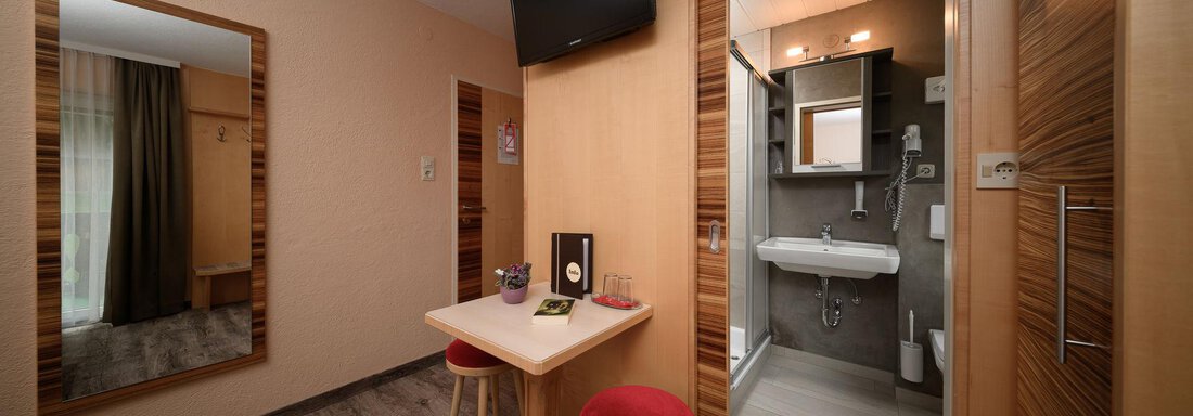 A modern room with a small table and two red chairs. In the background, there is a bathroom and a large mirror on the wall. - Haus Sonnschein - Zwieselstein