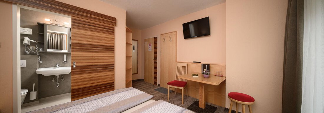 A modern room with two beds, a table, and a television. The bathroom is visible through a door, and the room has bright colors. - Haus Sonnschein - Zwieselstein