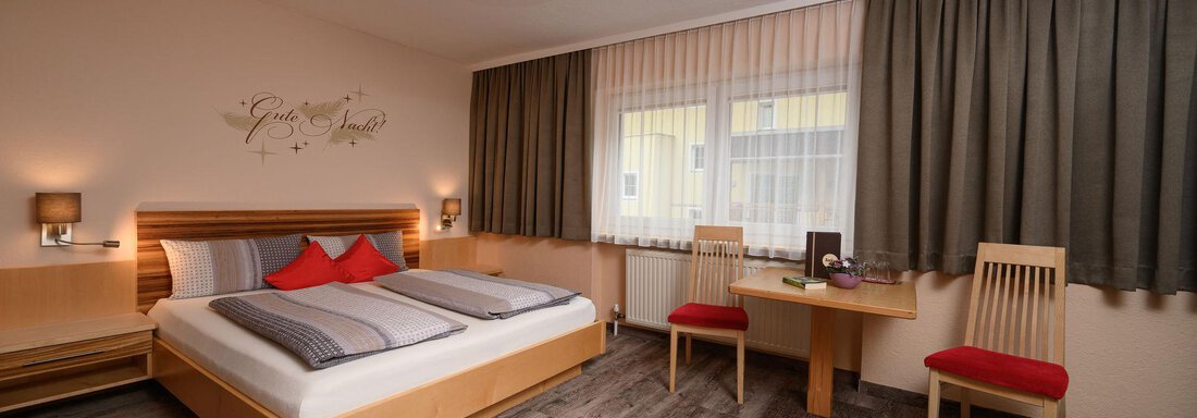 Doppelzimmer Komfort A modern hotel room with a large bed and cozy red pillows. There is a table with two chairs and large windows that let in natural light. - Haus Sonnschein - Zwieselstein