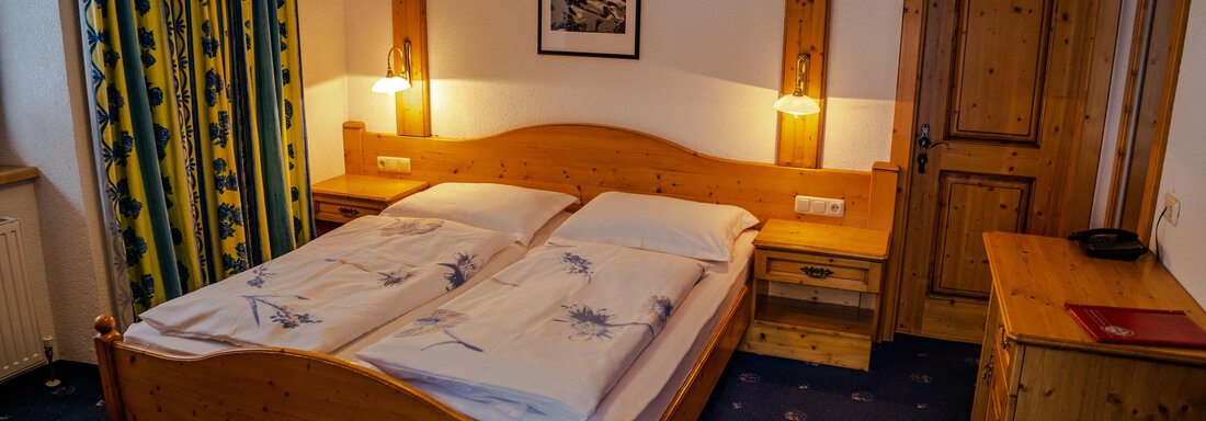 DZ2 A cozy bedroom with a wooden double bed and blue carpet. The walls are light and there are curtains with a colorful pattern. - Hotel Gasthof Zwieselstein - Zwieselstein