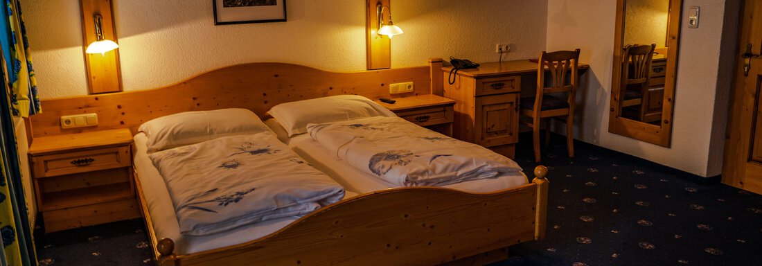 DZ3 A cozy room with two wooden single beds and warm light. There is a table, a chair, and a large mirror. - Hotel Gasthof Zwieselstein - Zwieselstein