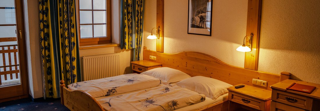DZ4 A cozy bedroom with a wooden double bed and two nightstands. Large windows with colorful curtains create a bright, inviting atmosphere. - Hotel Gasthof Zwieselstein - Zwieselstein