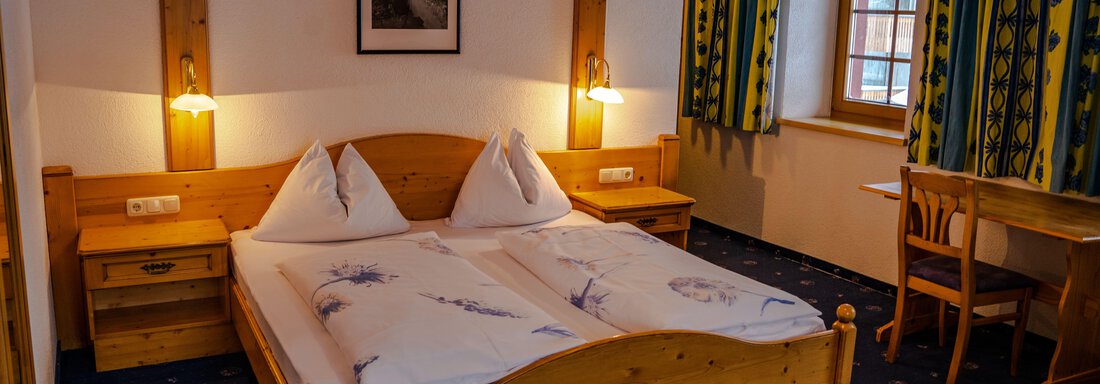 A cozy bedroom with two wooden beds, a beautiful wooden dresser, and warm light. The windows are adorned with colorful curtains, and there is a small table in the room. - Hotel Gasthof Zwieselstein - Zwieselstein
