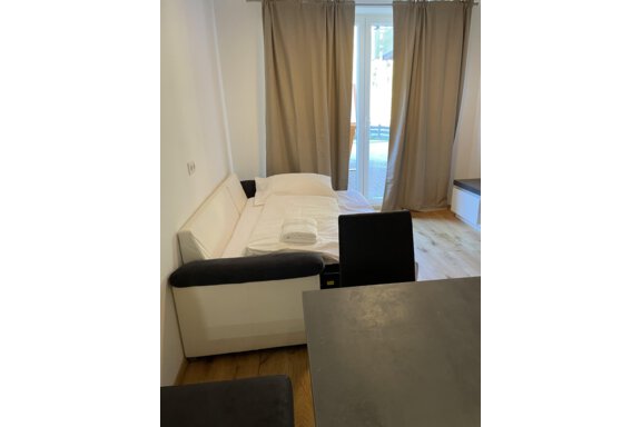 A simple room with a white sofa and a table. Light curtains hang in front of a window. - International House Sölden - Zwieselstein