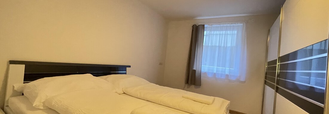 A bright bedroom with a double bed and fresh bed linens.  
A large wardrobe is against the wall and a window lets in daylight. - International House Sölden - Zwieselstein