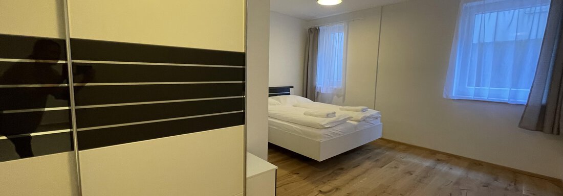 A modern bedroom with a white bed and a large wardrobe. The floor is made of wood and there are large windows that let in a lot of light. - International House Sölden - Zwieselstein