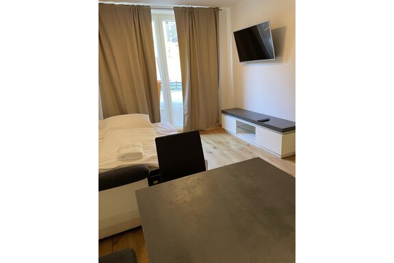 A simple room with a bed, a table, and a television.  
The windows are covered with curtains and provide access to a balcony. - International House Sölden - Zwieselstein