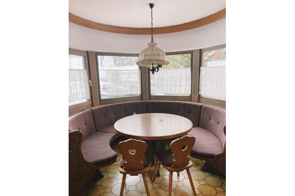 A cozy dining area with a round table and cushioned bench. Generous windows provide plenty of natural light. - Landhaus Rosenhof - Zwieselstein