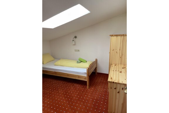 A simple bedroom with a single bed and a wooden dresser. The walls are white and the floor has a red carpet. - Landhaus Rosenhof - Zwieselstein