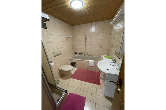 A bathroom with a bathtub and a shower. The floor is tiled and there are pink mats on the floor. - Landhaus Rosenhof - Zwieselstein
