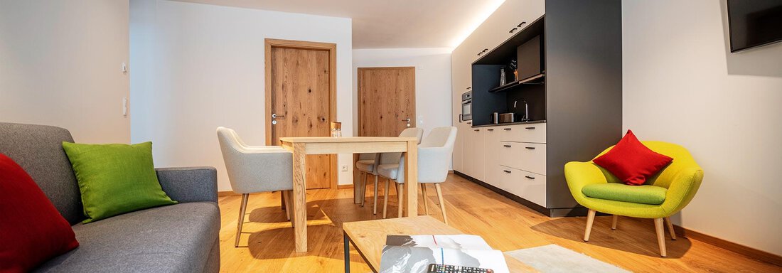 A modern living room with a gray sofa, a wooden table, and colorful chairs. The walls are light, and there is a cozy atmosphere. - Zwieselstein-Apart De Luxe - Zwieselstein