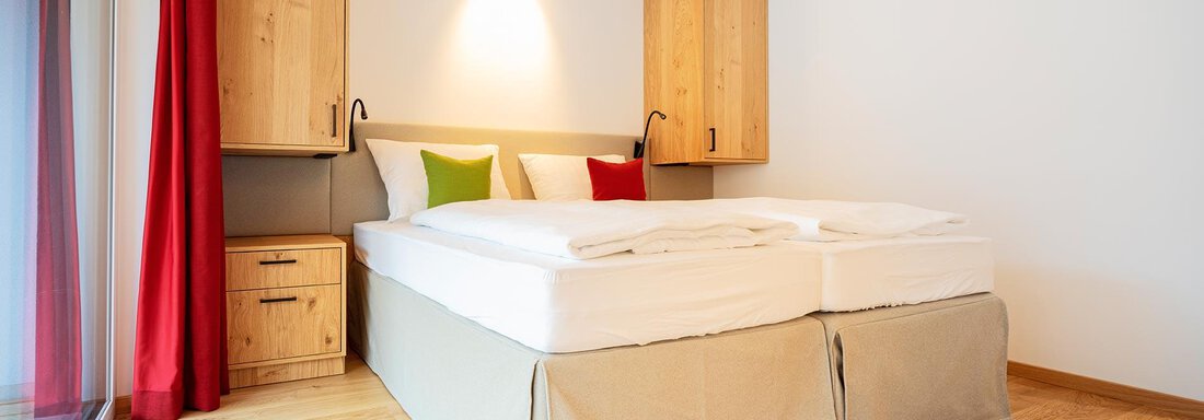 A modern bedroom with a double bed and wooden furniture. The walls are light and there are red curtains as well as cushions in various colors. - Zwieselstein-Apart De Luxe - Zwieselstein