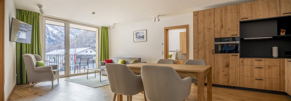 A modern living room with a dining area and an open kitchen. Large windows provide a view of the surroundings and plenty of daylight. - Zwieselstein-Apart De Luxe - Zwieselstein