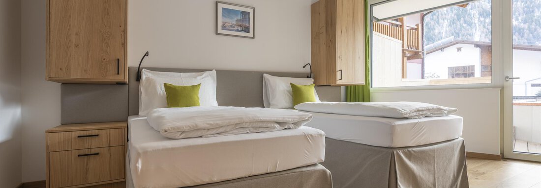 A cozy bedroom with two beds and wooden furniture. Large windows provide plenty of daylight and a view of the surroundings. - Zwieselstein-Apart De Luxe - Zwieselstein