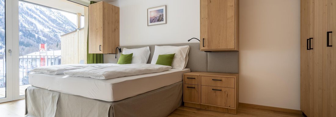 A modern bedroom with a large bed and wooden cabinets. A beautiful view of the mountains is visible through the windows. - Zwieselstein-Apart De Luxe - Zwieselstein