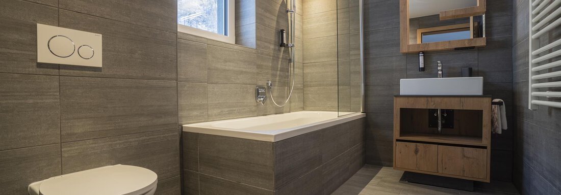 A modern bathroom with gray tiles and wood accents. It features a bathtub, a shower, and a stylish sink. - Zwieselstein-Apart De Luxe - Zwieselstein