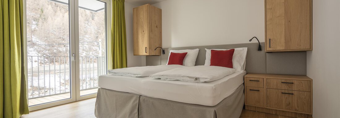 A modern bedroom with a large bed and wooden shelves. The windows provide plenty of daylight and a view of nature. - Zwieselstein-Apart De Luxe - Zwieselstein